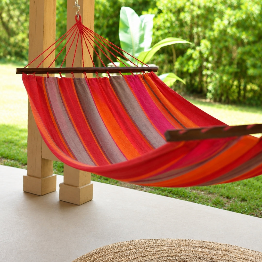 A striped fabric hammock in red, orange, pink, suspended between wooden posts on a patio, with a green garden background.