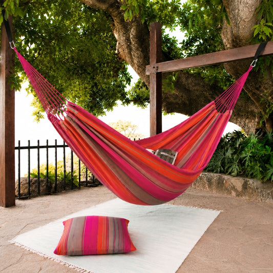 A striped hammock in pink, orange, red, and brown hangs between wooden posts under a large tree on a patio, with a matching striped pillow on a white rug below.