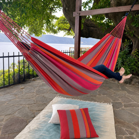 A striped canvas hammock in red, orange, pink, and gray is suspended between two trees on a stone patio. A person's legs and feet are stretched out inside the hammock.