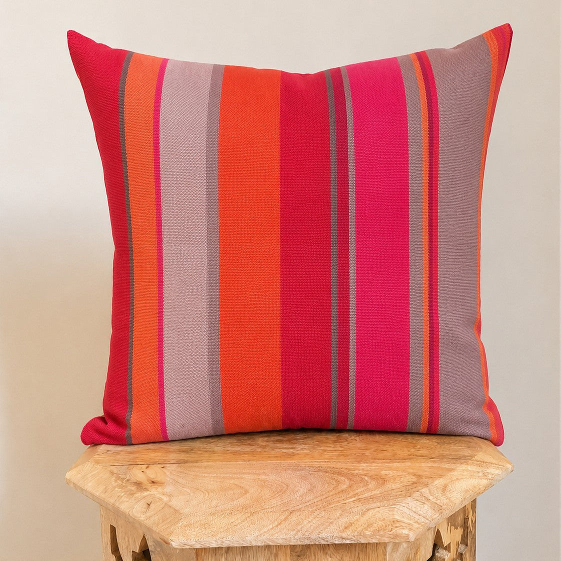 A square throw pillow with vertical stripes in red, orange, pink, and gray, resting on a carved wooden stool against a plain beige wall.