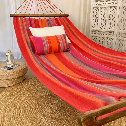 A colorful striped fabric hammock in shades of red, orange, pink, and purple, suspended over a woven jute floor mat. The hammock is styled with two decorative pillows, one white and one striped in pink, orange, and red, in a bohemian-style room setting with a carved white screen and a small woven stool.
