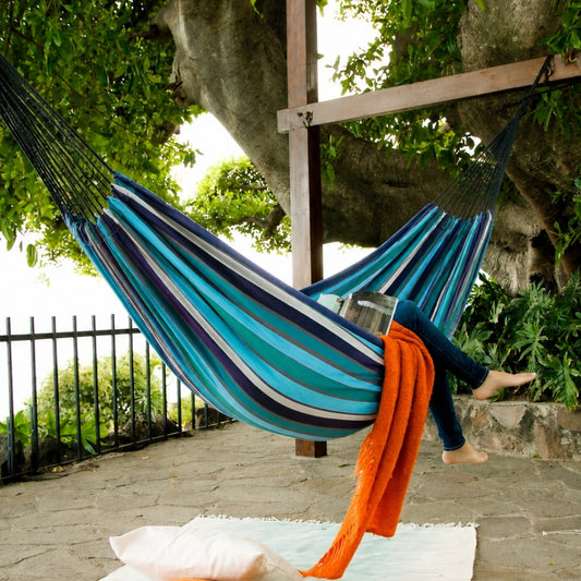 A striped outdoor hammock in shades of turquoise blue, teal, navy, and white, suspended between a wooden post and tree branches. A person's legs are stretched out in the hammock, with an orange throw blanket draped over the side.