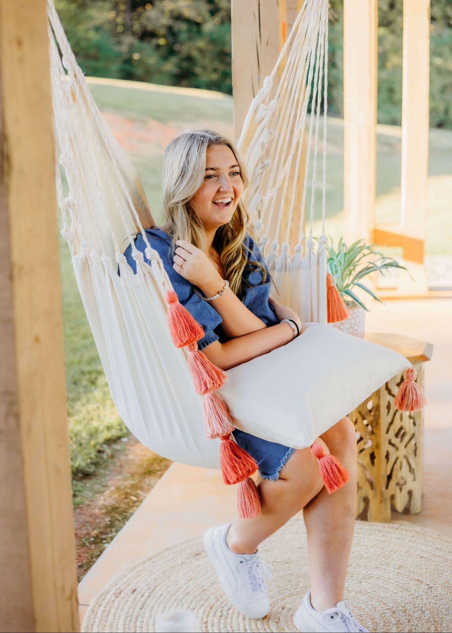 A woman sitting in a white macrame hammock with coral pink tassel accents on a wooden porch. 