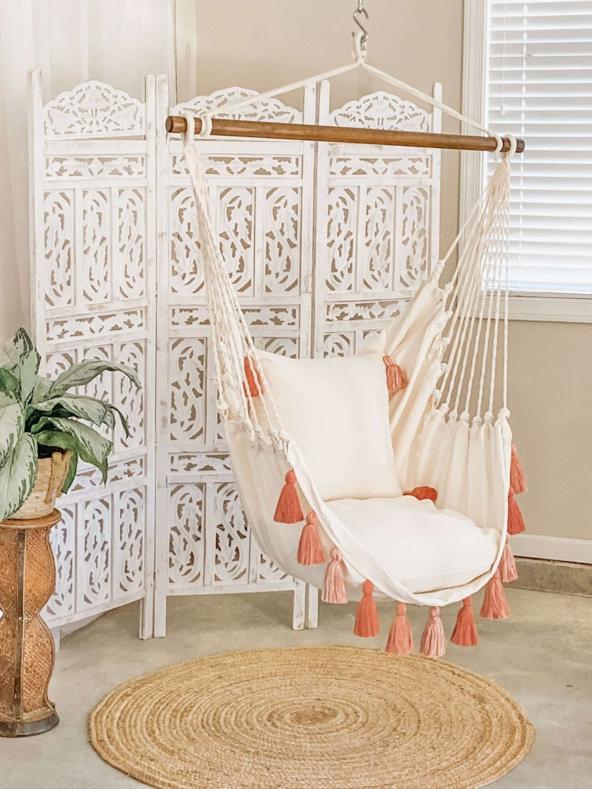 A white fabric hanging chair with coral pink tassel accents and a matching cushion, suspended from a wooden spreader bar. A round woven rug is on the floor.