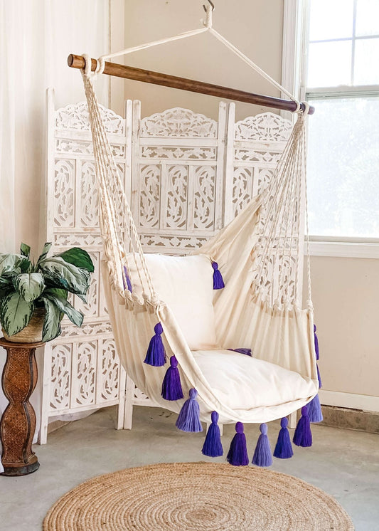 A cream fabric hanging swing chair with white cushions and purple tassels, suspended from a wooden bar in a bright room with a white decorative screen, window, plant, and woven rug.
